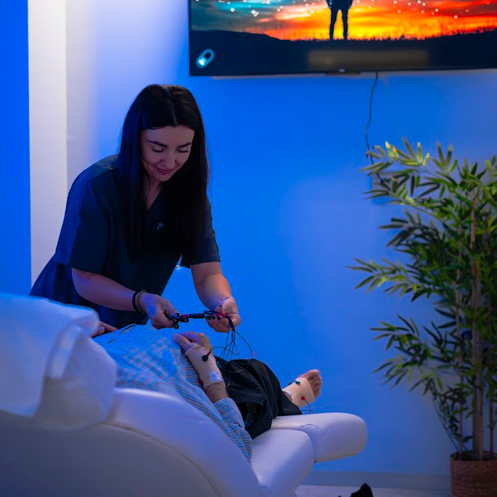Profesional de la salud aplicando un tratamiento de neuromodulación no invasiva a un paciente en una sala con iluminación relajante y pantalla con imagen estrellada.