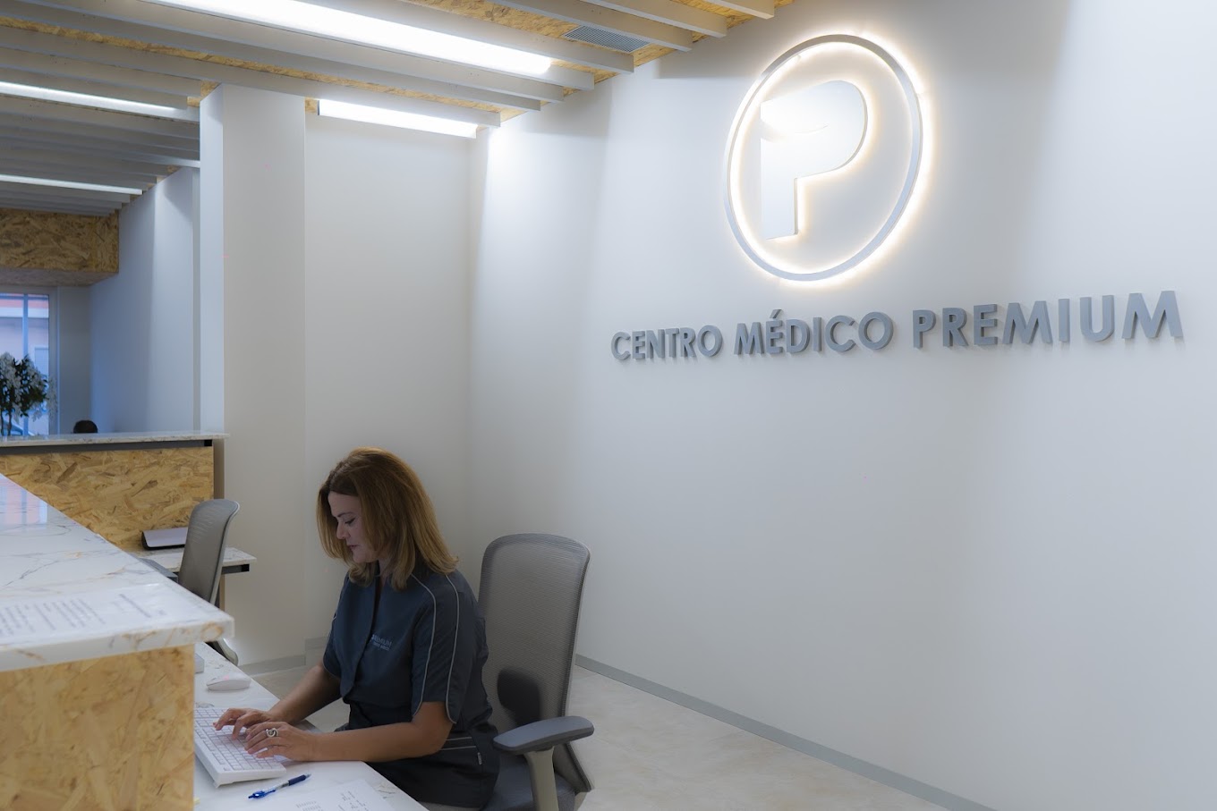 Recepcionista trabajando en el mostrador de Premium Centro Médico, con el logotipo iluminado en la pared de fondo y diseño moderno en madera.